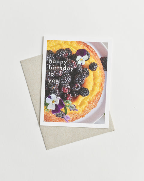 Photo greeting card of a round yellow cake topped with deep purple berries and flowers and white words “happy birthday to you!”